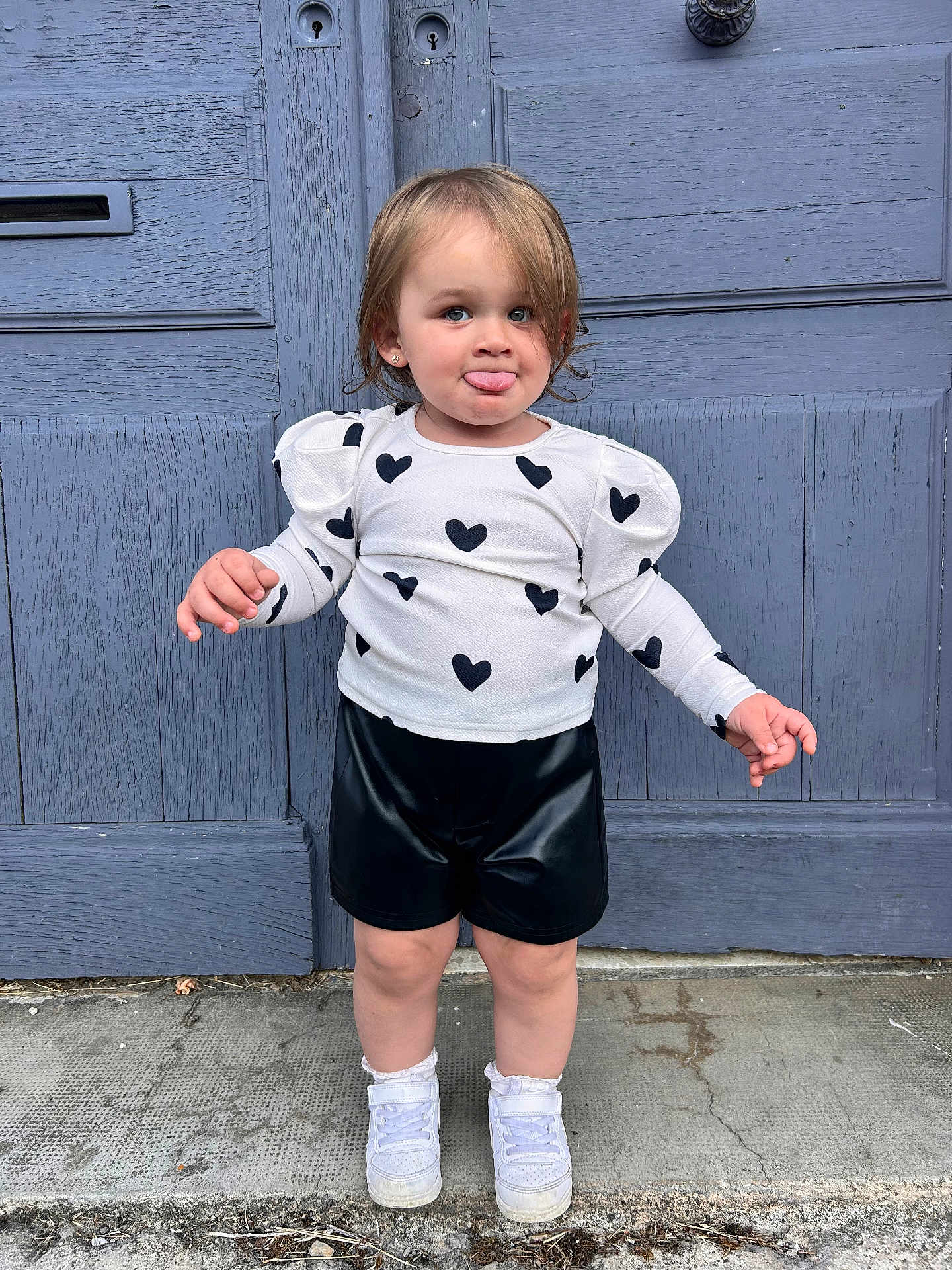 Julia participe au concours pour gagner de l'argent avec cette photo : toddler, child, person, standing, tongue_out, heart_pattern, white_top, black_shorts, white_shoes, door, blue_door, outdoor, concrete, sidewalk, cute, playful, fashion, young_child, smiling, casual_clothing