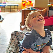 Corey is registered to the contest to win money with this photo: toddler, child, smiling, indoor, cushion, superhero_tshirt, happy, blond_hair, sitting, toy, home, floor, wooden_floor, furniture, playroom, casual_clothing, cute, portrait, young_child, bright