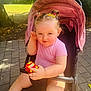 toddler, child, stroller, toy_car, outdoor, park, sunlight, hair_clips, pink_clothing, pavement, greenery, trees, smiling, cute, playful, baby, summer, daylight, person, seat