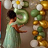child, girl, balloon, number_five, party, celebration, dress, green_dress, sandals, indoor, balloon_arch, wood_floor, curly_hair, decorations, birthday, flower_balloon, wall, standing, back_view, festive