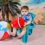 Ezra is registered to the contest to win money with this photo: toddler, child, plastic_chair, red_bucket, beach_ball, hawaiian_shirt, shorts, indoor, fake_palm_tree, beach_backdrop, serious_expression, barefoot, colorful_clothing, toy, summer_theme, portrait, playful, studio_photo, young_child, seated