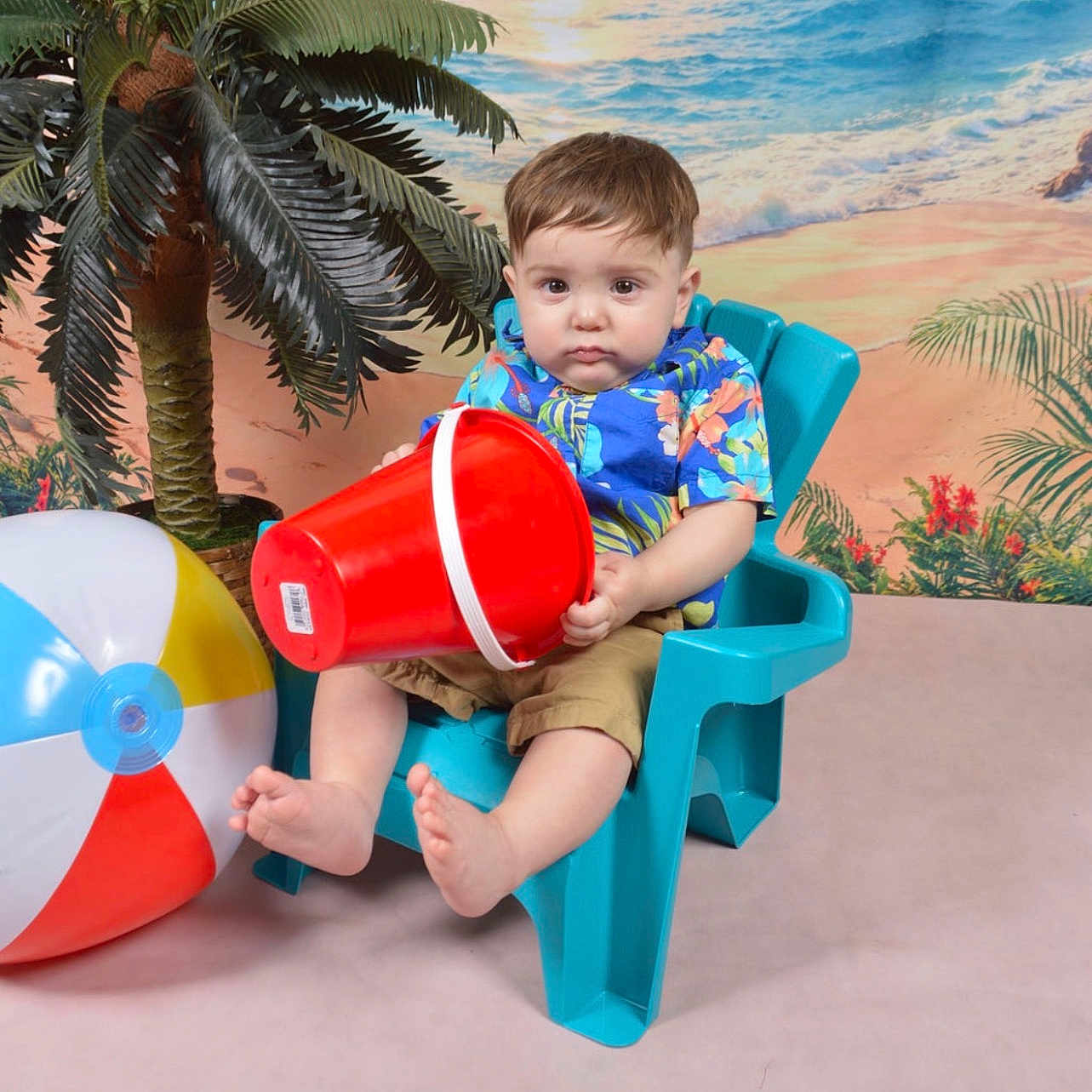 Ezra is registered to the contest to win money with this photo: toddler, child, plastic_chair, red_bucket, beach_ball, hawaiian_shirt, shorts, indoor, fake_palm_tree, beach_backdrop, serious_expression, barefoot, colorful_clothing, toy, summer_theme, portrait, playful, studio_photo, young_child, seated