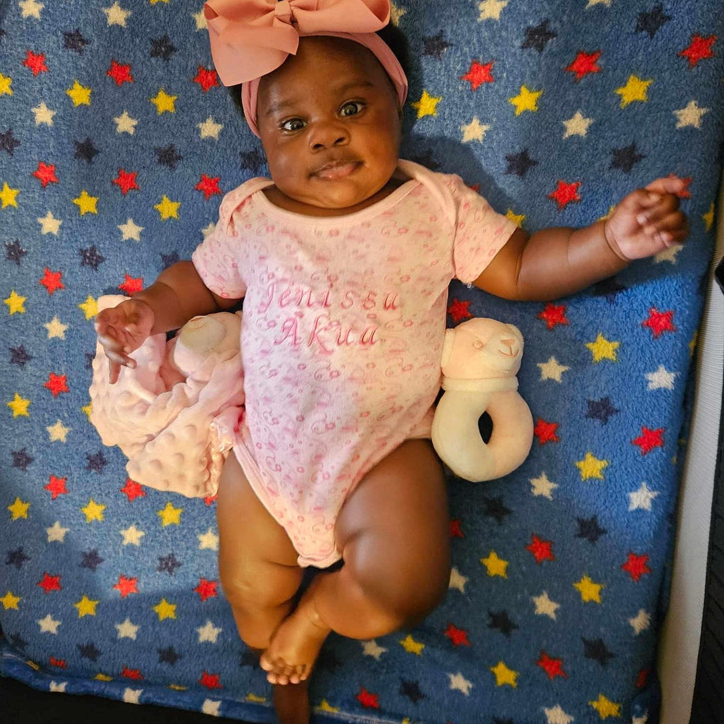 Jenissa joined the competition — help win amazing prizes! baby, blanket, child, comfort, cute, expression, face, foot, hand, headband, indoors, infant, lying_down, onesie, pink_bow, portrait, skin, smiling, soft_toy, star_pattern