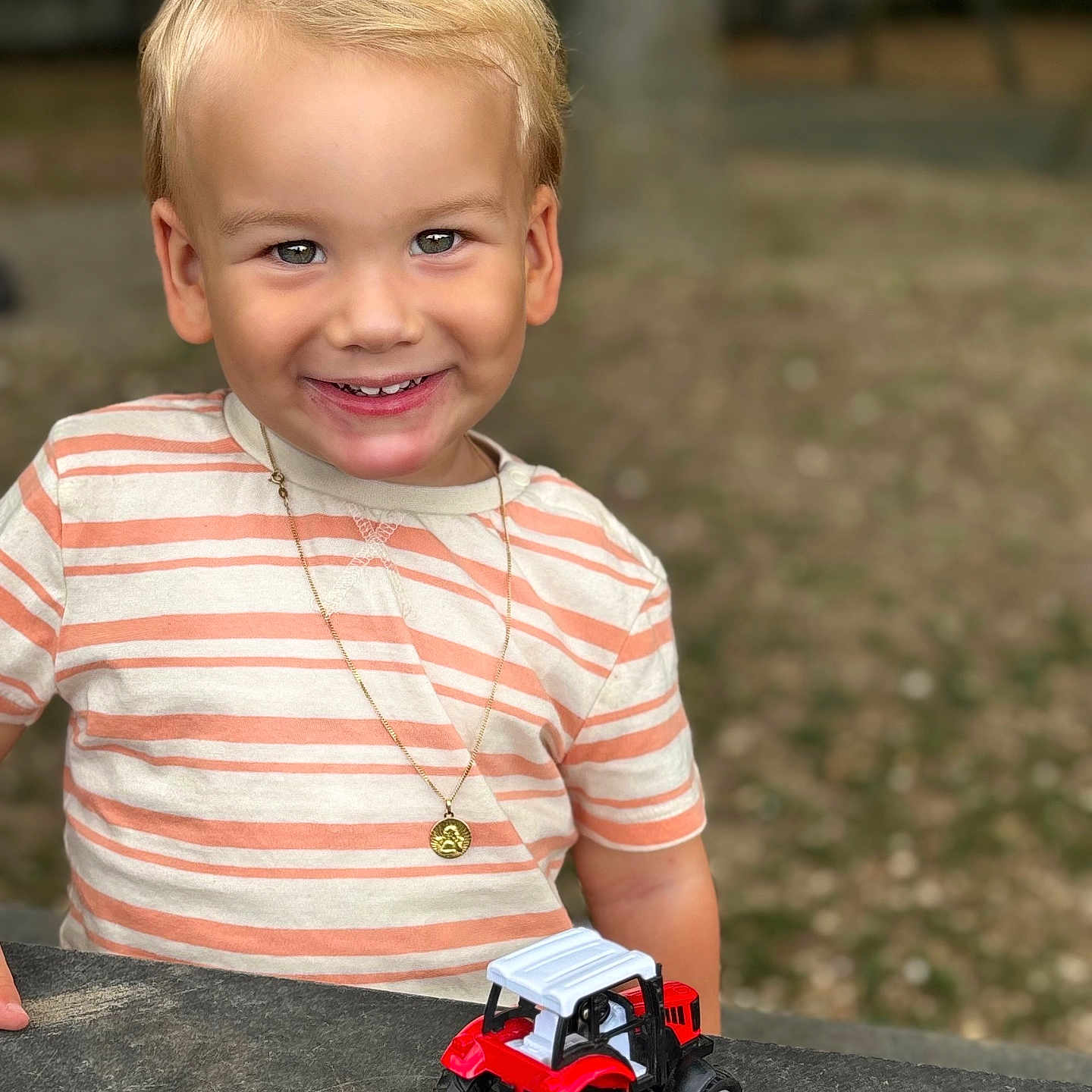 Côme participe au concours pour gagner de l'argent avec cette photo : blonde_hair, boy, casual_clothing, child, closeup, daylight, face, fun, grass, happy, nature, necklace, outdoor, play, portrait, smiling, striped_shirt, table, toy_tractor, tree