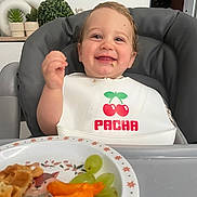 Ethan a rejoint le concours — aidez-le/la à gagner de superbes lots ! toddler, child, high_chair, bib, food, grapes, cheese_puffs, waffle, smiling, happy, indoor, plate, snack, face, person, home, decor, green_plants, vase, meal