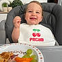 toddler, child, high_chair, bib, food, grapes, cheese_puffs, waffle, smiling, happy, indoor, plate, snack, face, person, home, decor, green_plants, vase, meal