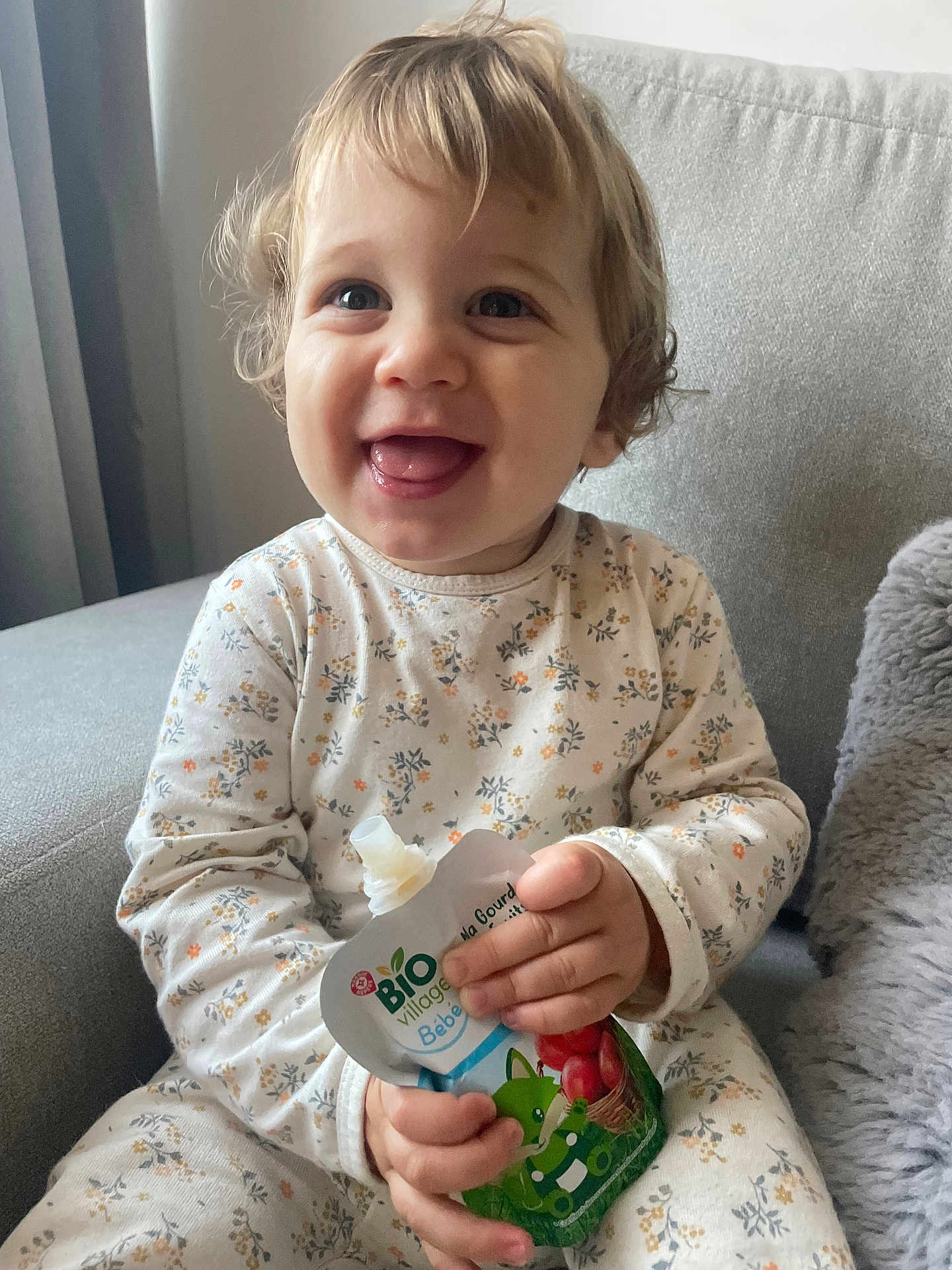 Ethan participe au concours pour gagner de l'argent avec cette photo : toddler, child, smiling, happy, baby_food, pouch, couch, curly_hair, blonde, onesie, floral_pattern, indoor, portrait, cute, young_child, seated, cozy, home, person, face