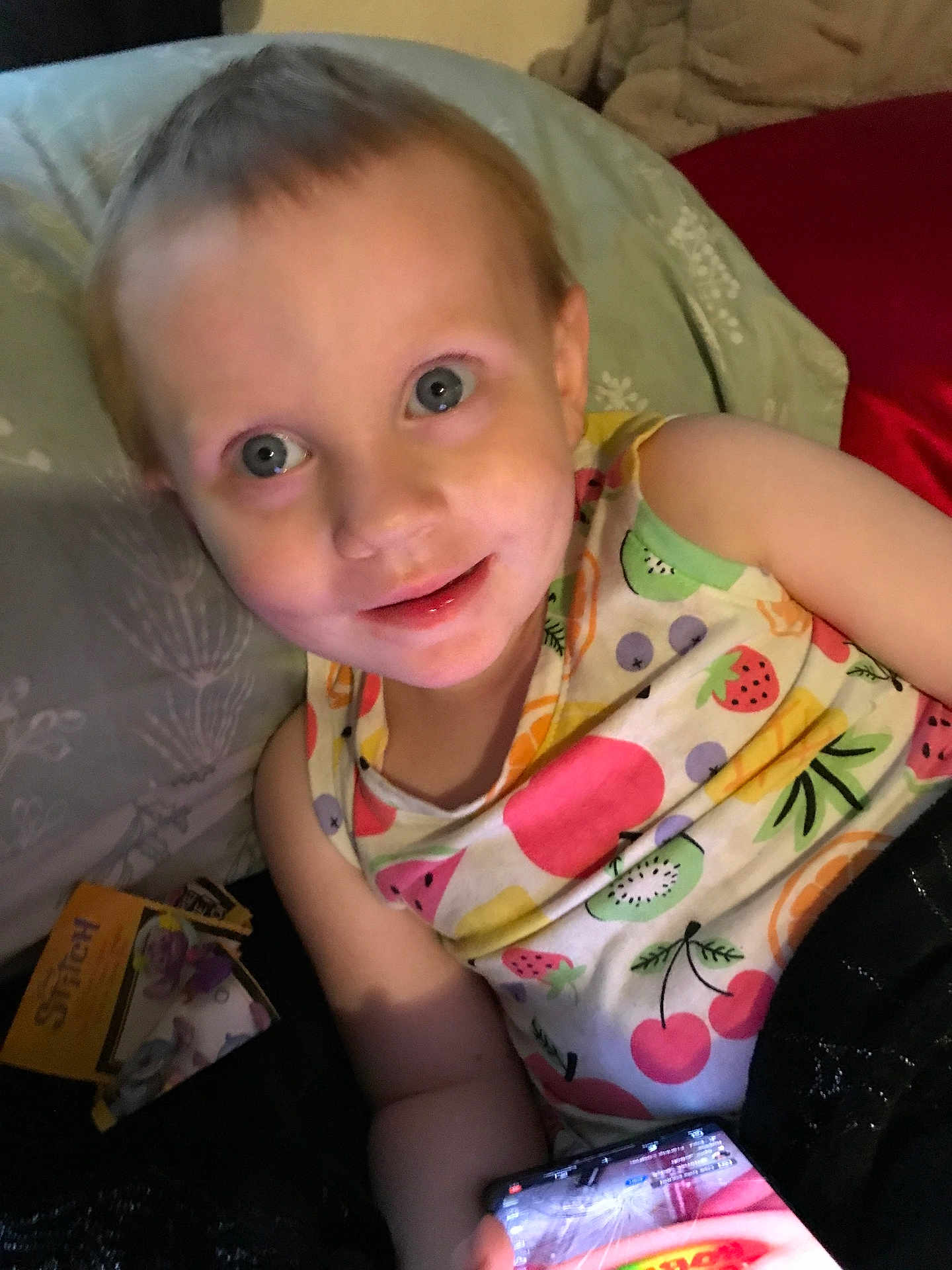 Summer is registered to the contest to win money with this photo: child, toddler, smiling, bed, pillow, blanket, colorful_clothing, fruit_pattern, smartphone, toy_package, indoor, person, face, happy, casual, young_child, relaxed, candid, portrait, blue_eyes