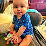 baby, infant, child, blue_onesie, toy, colorful_toy, playful, tongue_out, sitting, blanket, living_room, couch, pillow, plush, indoors, cute, chubby_cheeks, person, soft_lighting, happy