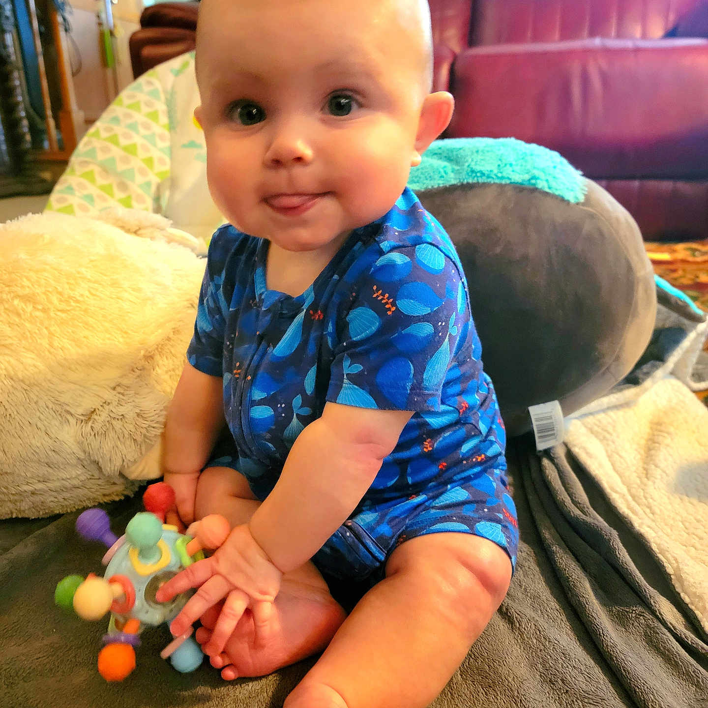 Silas is registered to the contest to win money with this photo: baby, blanket, blue_onesie, child, chubby_cheeks, colorful_toy, couch, cute, happy, indoors, infant, living_room, person, pillow, playful, plush, sitting, soft_lighting, tongue_out, toy