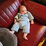 baby, infant, sleepsuit, blue_clothing, red_couch, leather_furniture, cushion, indoor, carpet, foot, hand, person, portrait, relaxing, seated, home_interior, soft_lighting, child, cute, cozy