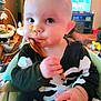 Silas joined the competition — help win amazing prizes! baby, child, high_chair, skeleton_outfit, food, spoon, messy_face, indoors, living_room, television, toy, furniture, cute, chubby_cheeks, person, seat, table, expression, hand, smile