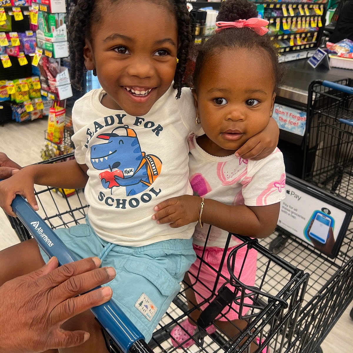 Khadija is registered to the contest to win money with this photo: background, bracelet, braids, child, children, cute, footwear, grocery_store, happy, person, pink_bow, retail, shopping, shopping_cart, shorts, smiling, snacks, store_aisle, supermarket, tshirt