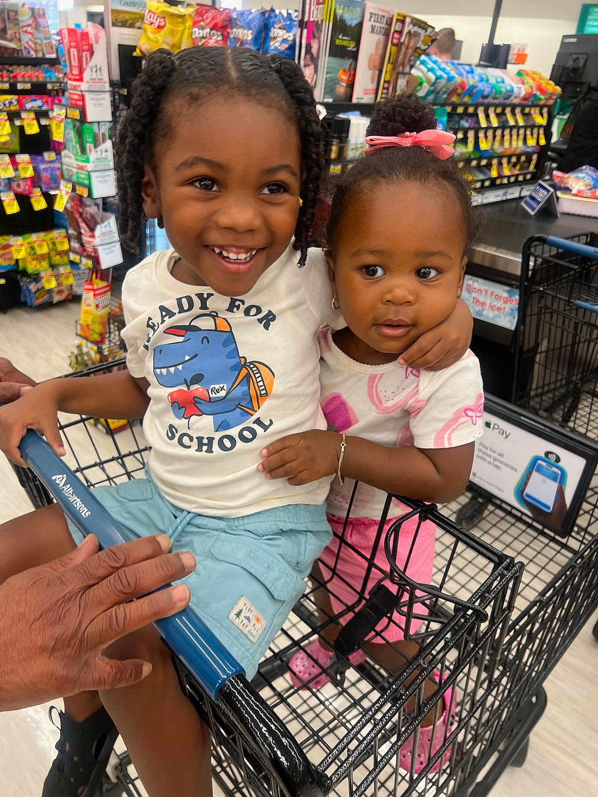 Khadija is registered to the contest to win money with this photo: background, bracelet, braids, child, children, cute, footwear, grocery_store, happy, person, pink_bow, retail, shopping, shopping_cart, shorts, smiling, snacks, store_aisle, supermarket, tshirt