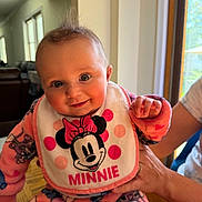 Victoria is registered to the contest to win money with this photo: baby, child, smiling, indoor, bib, minnie_mouse, pink_pajamas, person, hand, window, natural_light, cute, face, portrait, infant, clothing, happy, table, adult_hand, home