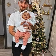 Deja is registered to the contest to win money with this photo: man, baby, child, christmas_tree, holiday_decorations, indoor, smiling, holding, festive, ornaments, lights, snowflake_decor, white_shirt, cap, pants, flooring, door, shelf, home, family