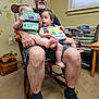 man, child, toddler, book, reading, chair, indoor, carpet, window, wall_decor, bookshelf, footwear, socks, shorts, smile, person, family, toy, room, wooden_furniture