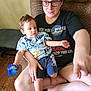 adult, child, toddler, glasses, toy, couch, birthday_badge, clothing, shorts, tshirt, wooden_floor, indoor, person, seated, holding, happy, home, casual, skin, hand