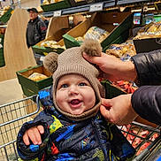 Joseph is registered to the contest to win money with this photo: baby, child, shopping_cart, hat, pom_pom, jacket, dinosaur_pattern, grocery_store, produce, potatoes, smiling, happy, person, hands, winter_clothing, indoor, retail, shopping, cute, face