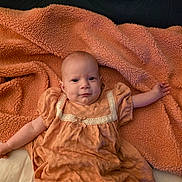 Summer is registered to the contest to win money with this photo: arm, baby, blanket, child, clothing, comfort, cute, dress, fabric, face, happy, head, indoor, infant, laying_down, orange, portrait, skin, smiling, soft_texture