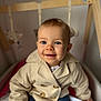 baby, child, smiling, blue_eyes, beige_coat, crib, toy, indoor, portrait, cute, infant, clothing, face, happy, person, young_child, seated, wooden_crib, soft_toys, cozy