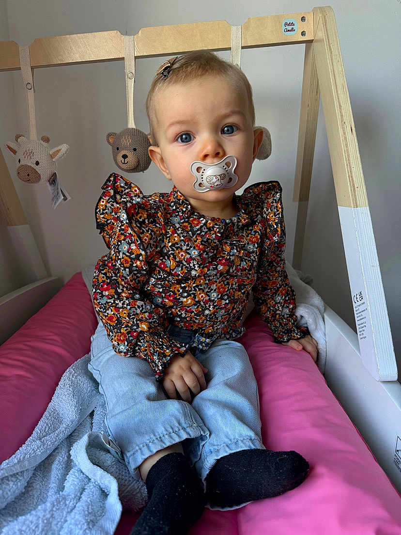 Ambre participe au concours pour gagner de l'argent avec cette photo : toddler, pacifier, blue_eyes, floral_top, jeans, black_socks, baby_gym, crocheted_toys, giraffe_toy, bear_toy, pink_cushion, blanket, wooden_frame, indoor, child, cute, sitting, portrait, baby, clothing