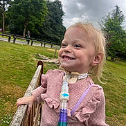 Harlow joined the competition — help win amazing prizes! child, smiling, outdoor, park, grass, trees, cloudy_sky, medical_device, tracheostomy_tube, fence, pink_sweater, person, happy, nature, healthcare, young_child, daylight, casual_clothing, cute, standing