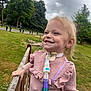 child, smiling, outdoor, park, grass, trees, cloudy_sky, medical_device, tracheostomy_tube, fence, pink_sweater, person, happy, nature, healthcare, young_child, daylight, casual_clothing, cute, standing