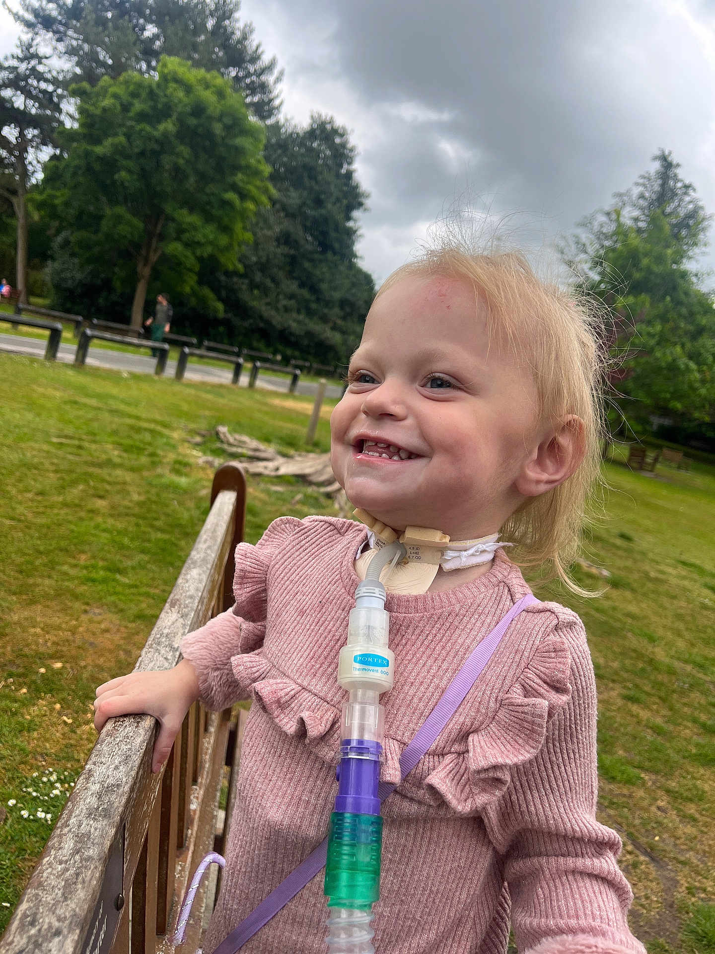 Harlow joined the competition — help win amazing prizes! child, smiling, outdoor, park, grass, trees, cloudy_sky, medical_device, tracheostomy_tube, fence, pink_sweater, person, happy, nature, healthcare, young_child, daylight, casual_clothing, cute, standing