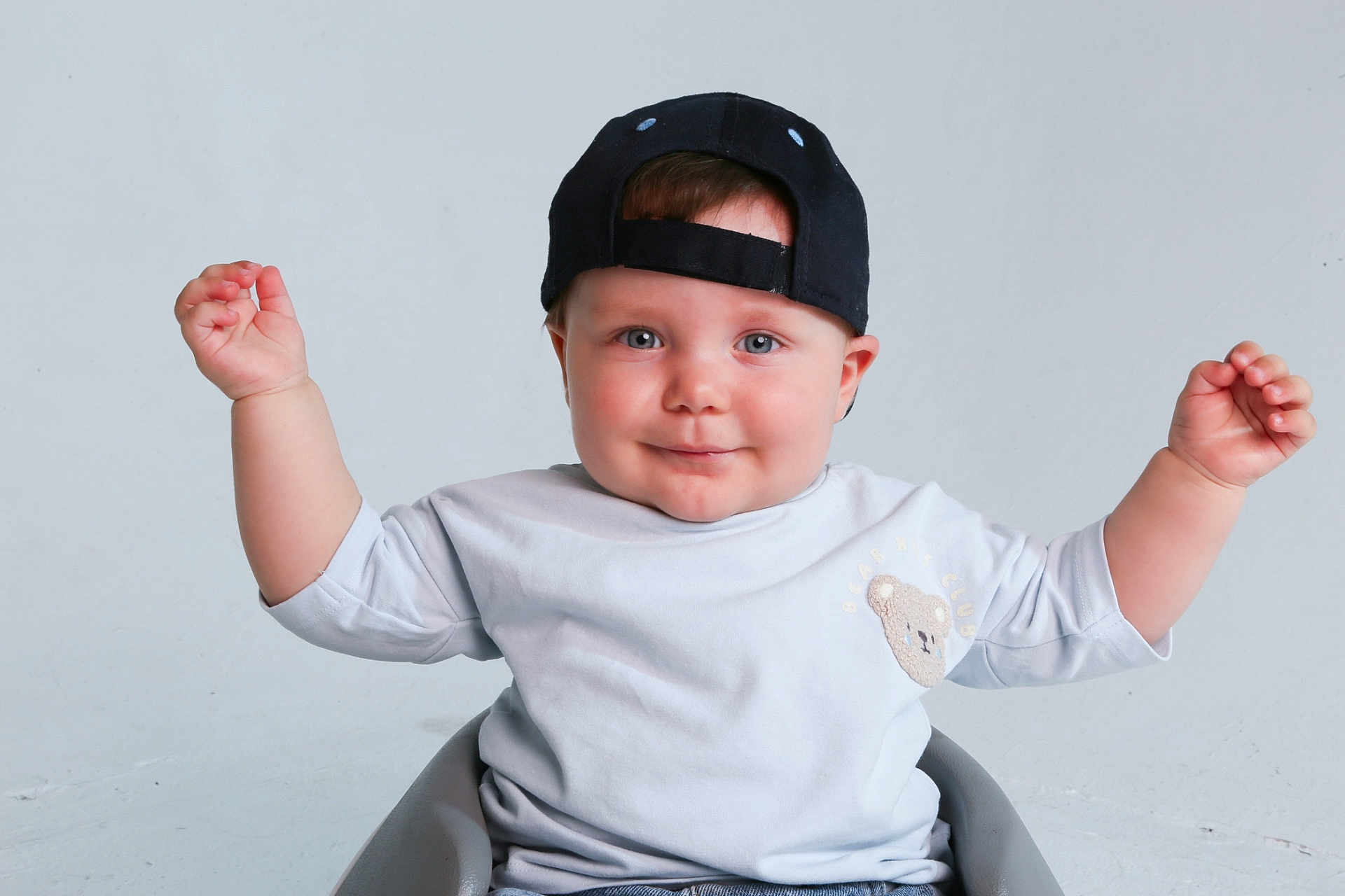 Mattias joined the competition — help win amazing prizes! baby, child, cap, smile, blue_eyes, teddy_bear_patch, light_shirt, sitting, raised_arms, happy, studio, minimalist_background, cute, portrait, infant, hands_up, casual_clothing, headwear, expression, indoor