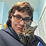 person, cat, glasses, indoor, selfie, tabby_cat, cuddle, face, hair, hoodie, earbuds, wall, door, light, eyewear, pet, comfort, companion, portrait, casual