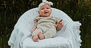 Emery is registered to the contest to win money with this photo: baby, infant, smiling, outdoor, nature, greenery, chair, blanket, headband, knitted_outfit, grass, happy, cute, portrait, sitting, child, person, daylight, forest, leaves