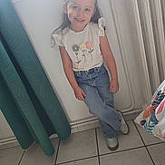 Angèle a rejoint le concours — aidez-le/la à gagner de superbes lots ! child, girl, smiling, indoor, white_wall, curtain, jeans, shirt, flowers, braids, shoes, tile_floor, radiator, pillow, casual_pose, happy, portrait, young, natural_light, home