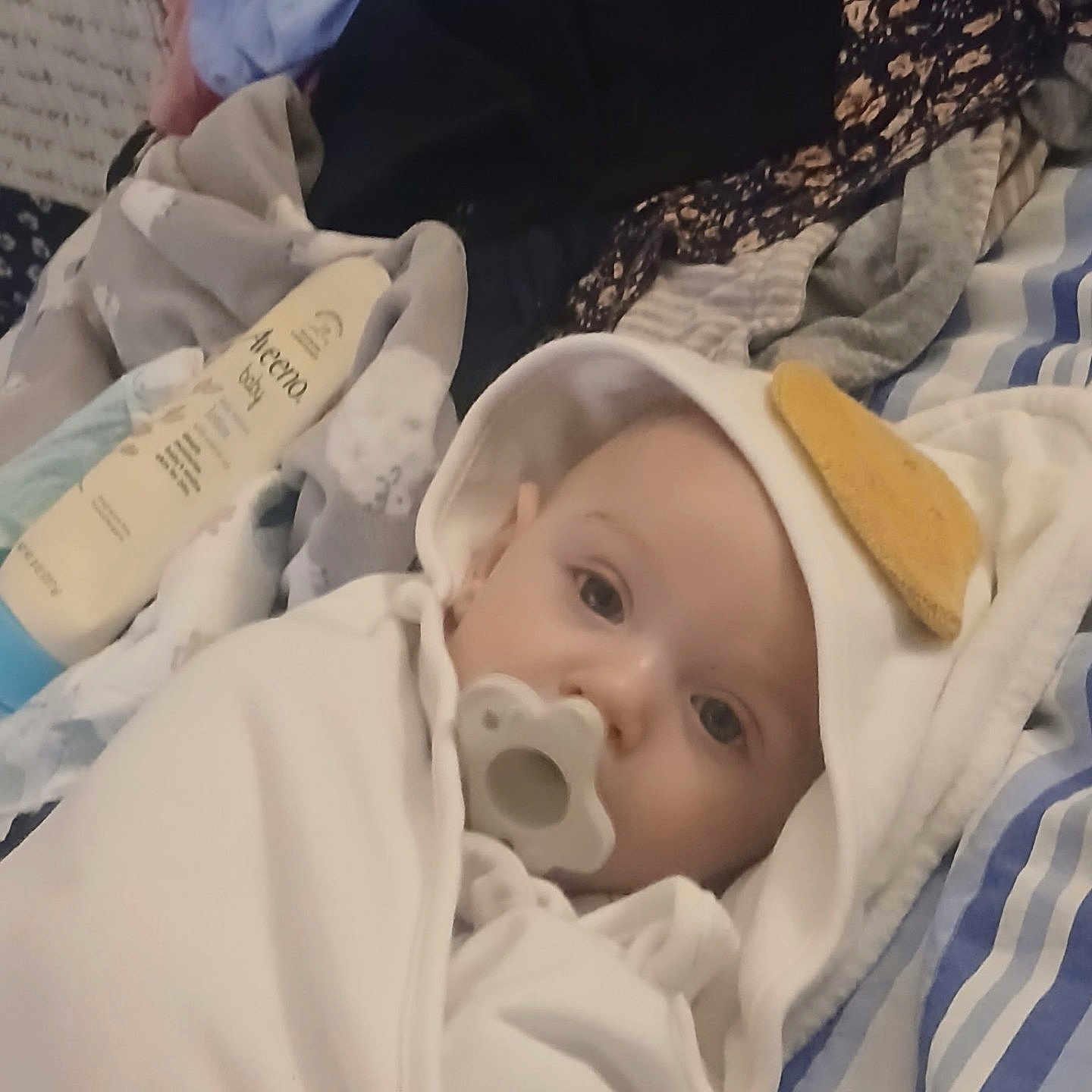 Acesen is registered to the contest to win money with this photo: baby, bed, blanket, child, closeup, clothes, comfort, cute, hooded_clothing, indoor, infant, lotion_bottle, pacifier, person, portrait, resting, sleepwear, soft_toys, toddler, white_clothing