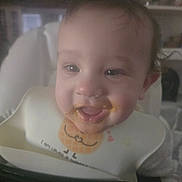Samson is registered to the contest to win money with this photo: baby, child, high_chair, bib, messy_face, food, smile, happy, indoor, person, cute, infant, face, hand, clothing, home, furniture, young_child, feeding, portrait