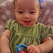 Eden is registered to the contest to win money with this photo: baby, child, smiling, toy, colorful, green_dress, sitting, indoor, cushion, polka_dot, hand, face, happy, curly_hair, infant, soft_texture, background, television, blurred, person
