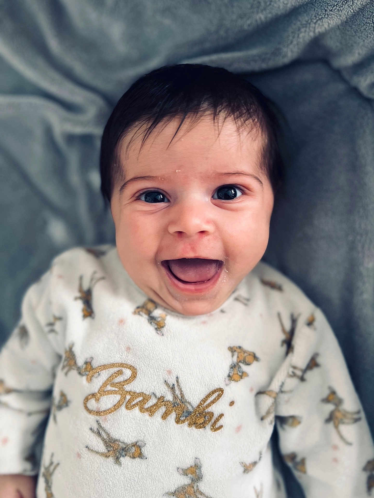 Mïa participe au concours pour gagner de l'argent avec cette photo : baby, infant, smile, laughing, close_up, portrait, onesie, text_on_clothing, bambi_design, dark_hair, big_eyes, open_mouth, cheeks, blanket, soft_texture, cozy, bed, newborn, adorable, candid