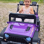 Aurora is registered to the contest to win money with this photo: balloons, child, daylight, dress, fun, gravel, greenery, happy, headwear, joy, outdoor, parked, play, playtime, purple, small_vehicle, smiling, toddler, toy_car, vehicle