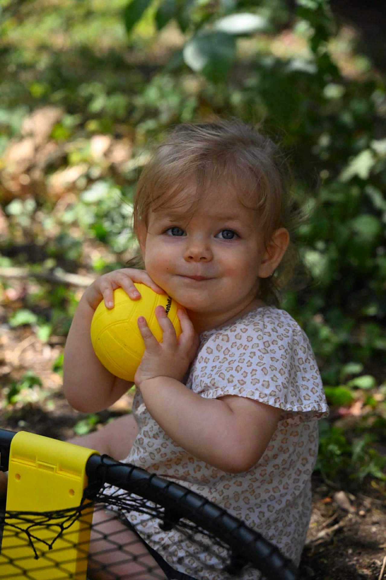 Sofia a rejoint le concours — aidez-le/la à gagner de superbes lots ! toddler, child, yellow_ball, outdoor, greenery, smiling, short_sleeve_shirt, patterned_clothing, grass, nature, play, fun, baby, person, face, hands, summer, sunlight, happy, cute