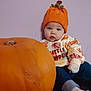 Ivy is registered to the contest to win money with this photo: baby, child, pumpkin, orange_hat, sweater, cute, indoor, portrait, curious, seated, purple_wall, clothing, infant, fall, autumn, holiday, seasonal, face, headwear, smile