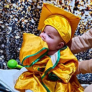 Ivy joined the competition — help win amazing prizes! baby, child, graduation_gown, graduation_cap, yellow_clothing, blanket, indoor, person, cute, chewing, sitting, soft_toy, supporting_hand, floor, celebration, portrait, infant, colorful_background, fabric, smiling