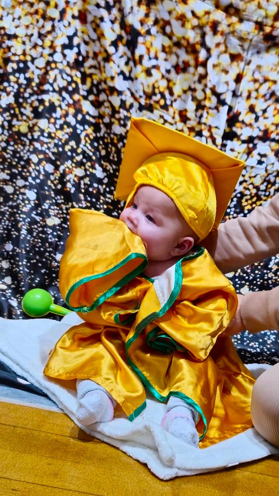 Ivy joined the competition — help win amazing prizes! baby, child, graduation_gown, graduation_cap, yellow_clothing, blanket, indoor, person, cute, chewing, sitting, soft_toy, supporting_hand, floor, celebration, portrait, infant, colorful_background, fabric, smiling