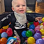 baby, child, ball_pit, colorful_balls, toys, skeleton_outfit, smiling, indoor, window, daylight, happy, playtime, infant, clothing, bib, face, person, home, furniture, cushion
