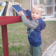 Kenzo a rejoint le concours — aidez-le/la à gagner de superbes lots ! blond_hair, blue_hoodie, books, bookshelf, building, child, curious, daylight, face, grass, green_pants, hand, outdoor, person, reading, sidewalk, sneakers, standing, window, young