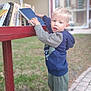 blond_hair, blue_hoodie, books, bookshelf, building, child, curious, daylight, face, grass, green_pants, hand, outdoor, person, reading, sidewalk, sneakers, standing, window, young