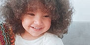 Sidra a rejoint le concours — aidez-le/la à gagner de superbes lots ! candid, casual, child, colorful, curly_hair, cute, face, hair, happy, indoor, joyful, patterned_fabric, person, portrait, sitting, skin, smiling, toddler, white_shirt, young_child
