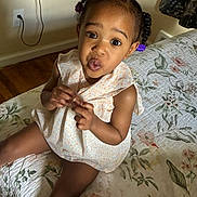Azaria is registered to the contest to win money with this photo: child, toddler, braided_hair, purple_hair_clips, dress, bed, bedspread, floral_pattern, indoor, wooden_floor, curious_expression, button, person, cute, young_child, seated, home, natural_light, playful, portrait
