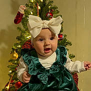 Zoe is registered to the contest to win money with this photo: baby, child, christmas_tree, holiday, festive, ornaments, lights, star, green_dress, white_headband, bow, socks, smiling, hands, indoors, decoration, celebration, person, happy, seasonal