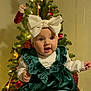 baby, child, christmas_tree, holiday, festive, ornaments, lights, star, green_dress, white_headband, bow, socks, smiling, hands, indoors, decoration, celebration, person, happy, seasonal