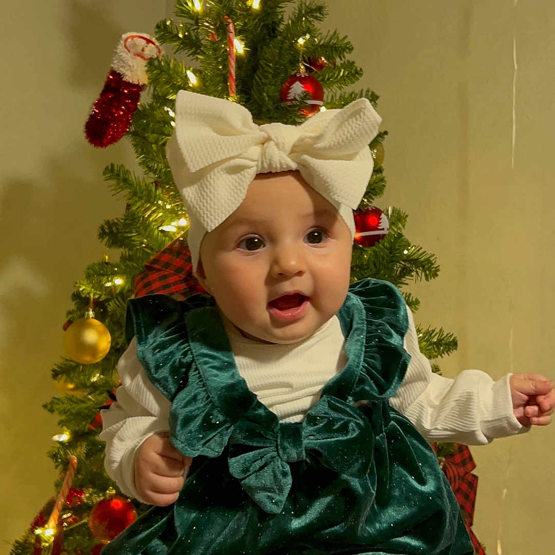 Zoe is registered to the contest to win money with this photo: baby, bow, celebration, child, christmas_tree, decoration, festive, green_dress, hands, happy, holiday, indoors, lights, ornaments, person, seasonal, smiling, socks, star, white_headband